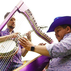 Larry Bellorín, a stout Hispanic man, sits and plays a magenta harp, wearing a purple cowboy hat and purple calico shirt. His bandmate Joe Troop, a white man, stands behind the harp and plays a banjo.