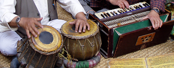 Tabla drums and a harmonium are being played by two Nepalese men sitting on a sisal rug in Khatmandu. Only the men’s hands are visible.