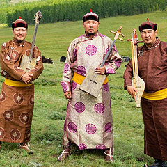 The three musicians of Alash Ensemble stand on a mountain field, holding traditional stringed instruments. They are all men of Asian appearance, all from Tuva in southern Siberia. They wear the garments of their culture: ankle-length coats of stiffened silk with bright gold sashes, caps with fur trim, and boots.