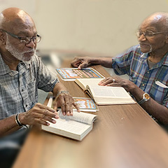Two Black men sit across from one another at a table, studying Bibles. The elderly man is Reverend Moses Jackson and the younger man (though old enough to have a gray beard) is his son Anthony.
