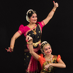 Rachita Nambiar and her daughter strike a pose as they perform a traditional dance from southern India. They are both smiling and holding their arms and fingers in specific, complex gestures. They are wearing silk saris in bright reds and gilts that contrast with the black background, and wear stylized makeup to accentuate facial expressions that are an important part of the dance.