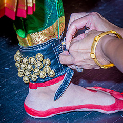 A woman’s hands buckle a band of small brass jingle-bells onto a dancer’s ankle. The dancer’s foot is rimmed in red paint, and she wears colorful silk leggings.