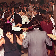 A very crowded room, with people holding hands as they dance in concentric rings. They are all wearing suits and nice dresses because they are attending a wedding reception.
