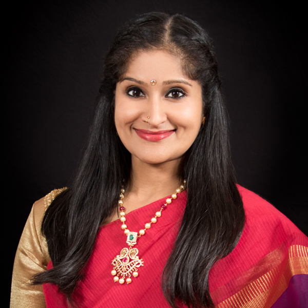A head-and-shoulders portrait of Rachita Nambiar. She appears to be in her thirties, with smooth dusky skin and long black straight glossy hair pulled away from her face and falling well past her shoulders.  She is smiling brightly; her dark eyes are wide open. She is wearing lipstick and eye makeup, and has a gold bindi (dot indicating she’s Hindu) between her eyebrows and a thin gold ring in her right nostril. Her sari is mostly cherry red, with gold on the right sleeve and purple on the left. She also wears a pendant necklace of gold beads and colored stones.