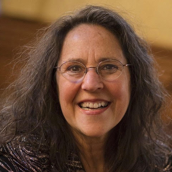 Susan Leviton smiles for a closeup portrait.  She has very wavy long hair, round glasses, fair skin, and is wearing a dark jacket with elaborate metallic embroidery.  She appears to be in her 50s.