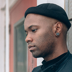 Carl “Buffalo” Nichols is seen in profile, wearing a black knit cap, a black shirt, and a large black disc earring. He is facing left, looking downward, seeming relaxed but serious and a little sad.