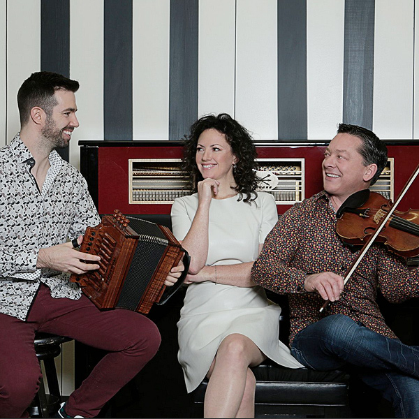 The three musicians of Cécilia, sitting in front of a piano: A young man holding an accordion, at left, a young woman who plays piano, and a man playing a fiddle (right). They are all looking at one another and grinning.