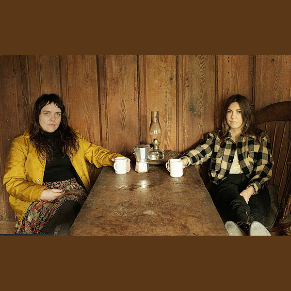 Mama's Broke: two young women sitting at a wooden table, facing the camera, with mugs and a teapot between them on the table