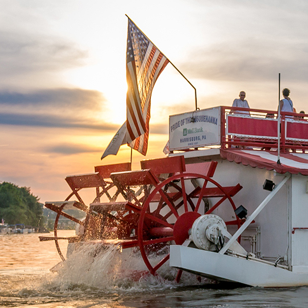 The Pride of the Susquehanna riverboat cruises on the river at sunset; this shot shows the two big red stern paddlewheels splashing water droplets, and passengers on the upper deck enjoying the view.