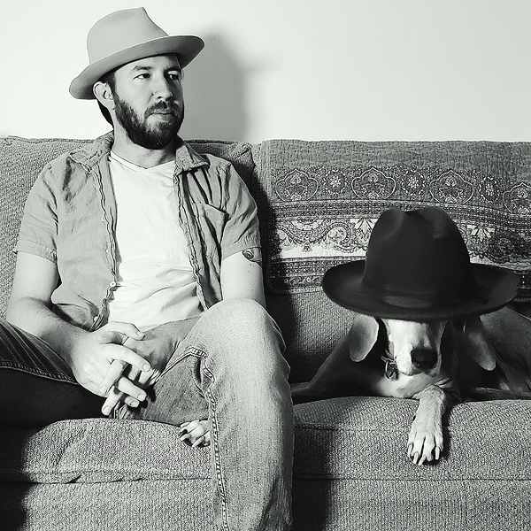 Zacharia Beaver, a bearded young man, sits on a couch wearing a cowboy hat. His bassett hound is beside him, with another cowboy hat over its head.