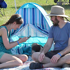 A young couple sits on a blanket, with a baby asleep behind them in a small colorful tent
