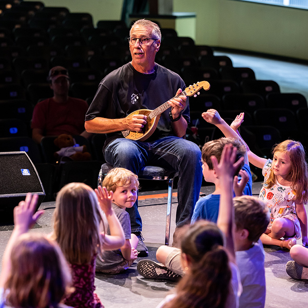 Lenny Tepsich sits on a chair, playing a small stringed instrument. Several young children sit on the floor in front of him; most are raising their hands and facing Lenny.  One younger boy, sitting right at Lenny's knee, has turned his head to look back at the camera; his expression is happy.