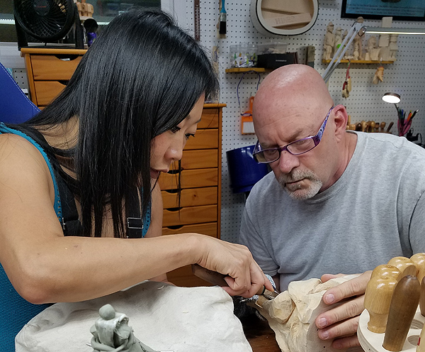 Aron Rook and Jim Hiser sit close together to work on a wood-carving. Aron is a young woman of Asian appearance, with straight black hair falling past her face. Jim is a portly man of middle age with a shaved head and short white goatee. He wears purple-framed reading glasses.