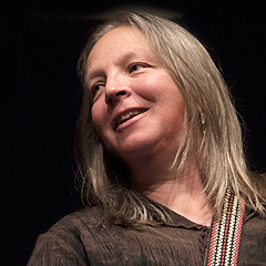 Barb Barton performing onstage, smiling and looking off to her right as if speaking to someone. She has a banjo on a colorful neckstrap but her hands are held together above it. She is wearing a dark brown shirt that blends in with the very dark background. Her grey-blonde hair is straight and the feathered ends fall on her shoulders.