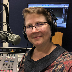 Debra D'Alessandro smiles serenely for a portrait in her radio studio.  She is wearing a headset over short blonde hair; she has a broad oval face and wears glasses. A broadcast microphone is poised in front of her on a mount. A mixing board with many tiny colorful controls is behind her at the bottom of the frame.  Further in the background, at the top of the frame, are two large computer monitors: one with software running, the other displaying a colorful wallpaper image.