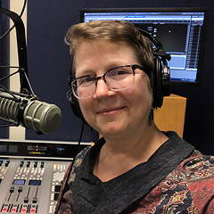 Debra D'Alessandro smiles serenely for a portrait in her radio studio. She is wearing a headset over short blonde hair; she has a broad oval face and wears glasses. A broadcast microphone is poised in front of her on a mount. A mixing board with many tiny colorful controls is behind her at the bottom of the frame. Further in the background, at the top of the frame, are two large computer monitors: one with software running, the other displaying a colorful wallpaper image.