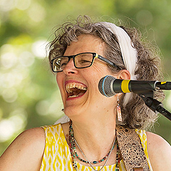 Jamie Anderson performing onstage, laughing and looking off to her right. She is playing a guitar and wearing a yellow-and-white dress with open shoulders. Her curly graying hair is held back from her face with a wide white hair band. She is wearing dark-framed glasses and four necklaces.