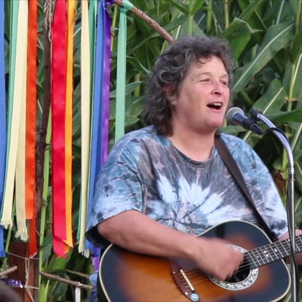Bobbi Carmitchell is playing her guitar and singing at an outdoor concert. Behind her is a field of corn; the corn plants are densely planted and much taller than she. Just in front of the corn to Bobbi's right, satin ribbons of various colors and widths hang from a curved branch, creating a colorful informal banner. She is wearing a tie-died shirt in shades of blue, and has dark curly shoulder-length hair that is starting to go gray.