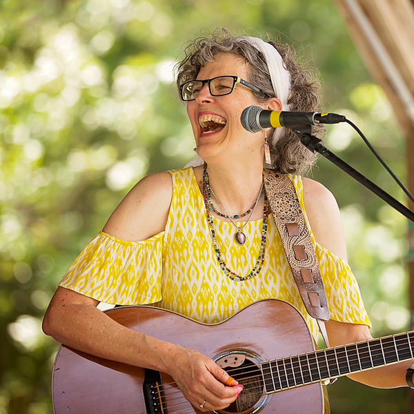 Jamie Anderson performing onstage, laughing and looking off to her right. She is playing a guitar and wearing a yellow-and-white dress with open shoulders. Her curly gray-blonde hair is held back from her face with a wide white hair band. She is wearing dark-framed glasses and four necklaces.