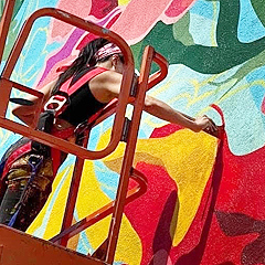 Aron Rook stands on a scaffold to paint a colorful mural on the side of a building