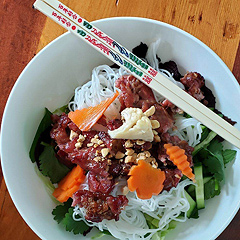 A plate of Vietnamese grilled pork. It is beautifully presented in a whitebowl with pretty chopsticks laid across the rim pf the bowl.  The bowl contains a layer of salad greens at the bottom, then a pile of thin white rice noodles, then the pork in red-brown sauce, then a garnish of decoratively-sliced carrot pieces, chopped peanuts, and cauliflower florets.