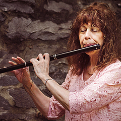 Lesl Harker plays a wooden flute. It is black wood with silver rings, and has no keys. She is a slim woman who appears to be in her 50s, with wavy auburn hair that falls over her shoulders and bangs that cover her forehead.  She is wearing a pink lace blouse, and her eyes are closed. A very rough stone wall is in the background.