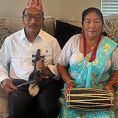 Two Nepali people sitting on a couch, holding instruments. They are Muskan's parents, and appear to be in their 60s. The man is holding a small stringed instrument with a bow. He is wearing a white dress shirt and a colorful almost-conical hat. The woman is holding a long skinny double-headed drum on her lap, with one hand on each drum head.  She is wearing a green silk sari and a tee shirt, with a multi-strand necklace of red beads.  She has dark hair, pulled back, and a small red bindi between her eyebrows.