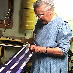 An elderly woman stands by a quilting frame, focusing on something small in her hands, wearing a dress and cap typical of Pennsylvania Mennonites.