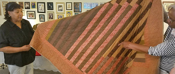 Cherry Lewis, a Black woman, holds a corner of a quilt she made, while another Black woman (mostly out of the frame at right) holds up an opposite corner and points to a detail on the quilt with her free hand.  The quilt is made of long, narrow horizontal strips of fabric, alternating dark and light.  Colors range from dark brown to peach; some strips are printed calico and some are solid color.