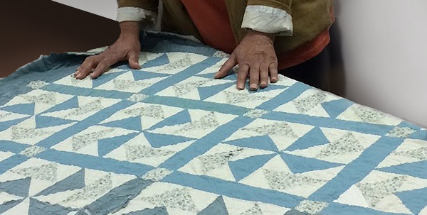 A quilt, a blue and white pinwheel pattern, is spread on a table. Narda LeCadre stands behind the table with her hands touching the quilt.  The photo is cropped so that only her hands and a portion of her body is visible; the quilt is the center of attention.