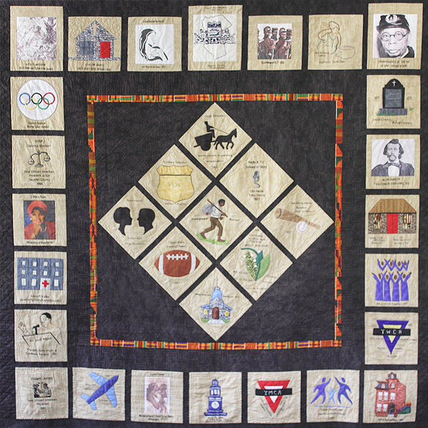 This quilt features tan ‘story squares’ with embroidered or appliqued pictures and lettering, each referring to a person or event, set on a dark background. Around the edge of the quilt are 24 story squares. A diamond of nine story squares occupies the center of the quilt, with a bright line of orange and green African print fabric separating the diamond from the outside squares.  Images on the story squares include a man walking with a bundle on his back, a football, a baseball and bat,  a horse-drawn wagon, a choir, primitive buildings, and photographs of people.