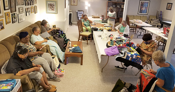 Nine members of the African American Quilters Gathering of Harrisburg meet in a large room. Four women sit on a long couch along the left-hand wall, sewing in their laps. The others sit at a long table running down the center, also working on quilts. Around them are sewing machines, neat piles of fabric,  and other quilting supplies.