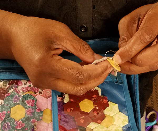 Closeup of a Black woman’s hands stitching fabric together. Each of the petals is a hexagon of colored calico, about an inch in diameter.  Each flower is made with six hexagon petals around a solid-yellow center hexagon.  A zippered bag with a clear plastic cover holds a dozen or so completed flowers.  A different calico is used for the petals of each flower; most feature a floral print.