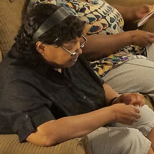 A Black woman sits on a couch, focusing on her hands which might be sewing something small. She is wearing glasses pushed down on her nose, and has curly hair held back with a shiny black band.