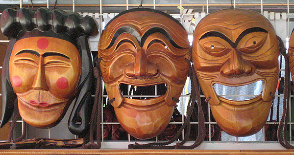 Three Talchum masks hang on a wire grid. The mask at left appears to be a woman, with shoulder-length hair and a horizontal roll of twined hair across the top. Quarter-sized red dots are painted on cheeks and forehead, and the center of the lips are painted the same red. Her eyes are closed and she seems to be smiling. The center mask is of a laughing man with prominent eyebrows and eyes squinched shut. The mask at right is of a bald man with wide-open eyes and wide-open semi-smiling mouth.