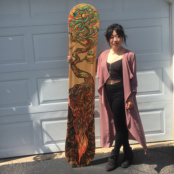 Aron stands in front of a white garage door, facing the camera, with one arm holding a decorated snowboard that’s a foot taller than she is.  It is decorated from top to bottom with a stylized tree. L eaves and fruit cover the top two feet of the board, roots and soil cover the bottom three feet, and a twisted trunk connects them.  Aron is a slim Asian woman, wearing a snug black crop top, black leggings, large black boots, and a knee-length jacket of thin, drapey, mauve fabric.