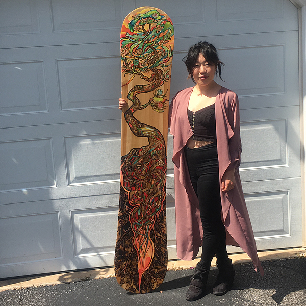 Aron stands in front of a white garage door, facing the camera, with one arm holding a decorated snowboard that’s a foot taller than she is. It is decorated from top to bottom with a stylized tree. L eaves and fruit cover the top two feet of the board, roots and soil cover the bottom three feet, and a twisted trunk connects them. Aron is a slim Asian woman, wearing a snug black crop top, black leggings, large black boots, and a knee-length jacket of thin, drapey, mauve fabric.