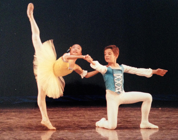 Aron Rook as a young girl, performing ballet wearing a yellow tutu with white tights. She is standing on one leg, on tiptoe but not en pointe, with her other leg stretched straight up toward the ceiling. The boy dancing with her is kneeling with his arms outstretched. Aron holds one of his hands, and rests her other hand resting on his shoulder. She gazes upward, well above the camera.
