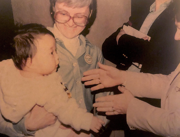 A woman with large-framed glasses holds 13-month-old Aron Rook on her hip with one arm, smiling down at her.  Another woman (mostly out of the image) holds out both hands as if to take the baby in her own arms.  The baby, who is looking at this new woman, wears a fluffy white hooded jacket; she has short black hair and Asian features. The woman reaching for her is wearing a pink suit jacket.  Behind these people, a third person in a dark blazer stands, with hands folded across their chest, holding some folded papers.