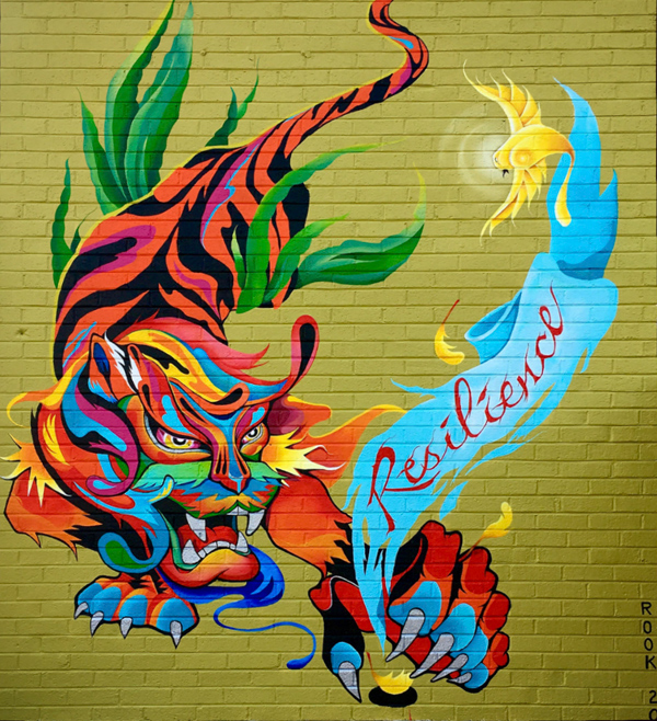 An Asian-inspired tiger reaches out a huge clawed paw to try to capture a bird.  But the tiger only gets a feather, as the bird flies up, trailing a ragged turquoise banner with the word ‘Resilience’ in red cursive letters.