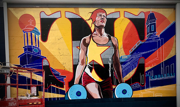 A mural on the side of a building, depicting a woman athlete with short red hair. She is holding dumbells in each hand, wearing red shorts and a gray tank shirt with a large gold graphic star covering half her chest. Line drawings of urban buildings are to her left and right. Behind her, there are rays of gold sunlight, and a large orange disc over her shoulder on the right side.