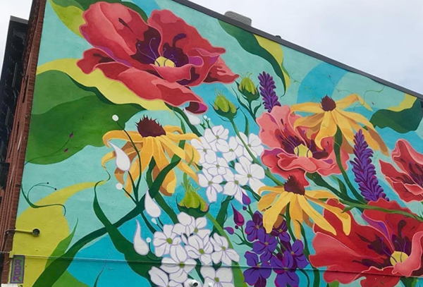 A mural on the side of a building, depicting gigantic flowers including red poppies, yellow coneflowers, clusters of white and purple phlox, and leaves in many shades of green.