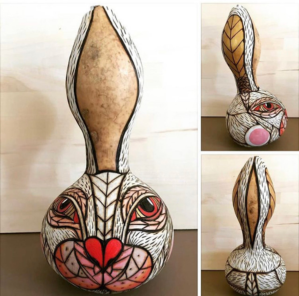 A gourd that has been decorated (with wood-burning) as a stylized rabbit. The round part of the gourd is its face -- with a heart-shaped nose, rather human eyes, and hash-marks indicating fur. The rabbit’s ears go up the sides of the gourd’s tall neck