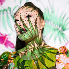 A soft-focus self-portrait photo showing Aron’s face and shoulders. Many small rhinestone gems decorate Aron’s face, and a large palm frond is painted across her neck, face and shoulders. The upper tips of the fronds almost reach her eyelids; the center stem curves up across her collarbone; and the lower tips of the fronds reach down across the neckline of her dress and merge with its green-and-peach floral print.  She is gazing dreamily at something above the camera; her left pupil is centered within the palm frond crossing that eye. Her black hair is pulled away from her face into a ponytail at the top of her head, and her jawline is silhouetted against the hair falling behind her right shoulder. Her lips are painted with dark red lipstick outlined in black, and are slightly parted to show her teeth. Behind her on the wall are more of the frondy leaves and some bright magenta patches that are probably flowers.