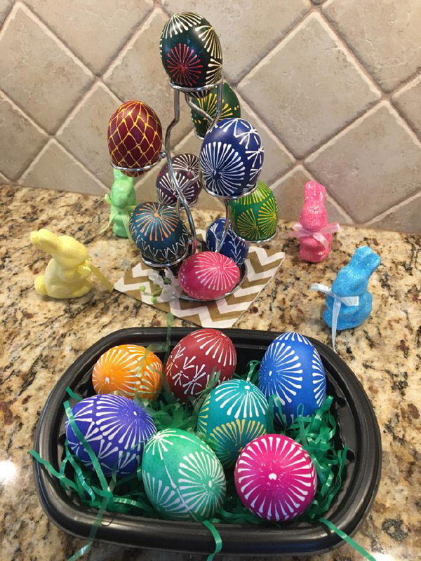 In the foreground: a black plastic bowl with a little plastic ‘Easter grass’ holds seven decorated eggs in red, green, blue, purple, orange and maroon. Most of the eggs feature variations on a ‘pinwheel’ pattern that resembles the spokes of a bicycle wheel radiating from a central dot. Most of the eggs have four of these pinwheels circling the egg, with smaller pinwheels on top and bottom. Behind the bowl, a wire ‘egg tree’ holds more decorated eggs in deeper colors, with some multicolored designs. Small bunny statues complete the display.