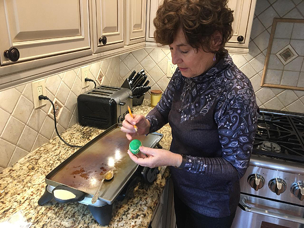 Barbara stands in the corner of her kitchen, with a hot griddle in front of her on the counter. On the griddle is her mother’s spoon, holding melted beeswax, its handle resting on the edge of the griddle. She is applying wax to a green-dyed egg, using her pencil-and-pin tool with her right hand. It looks like her right pinky is braced against the egg to steady her hand. She is looking down at the egg.