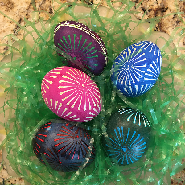 Five decorated eggs in pink, blue, green, red and maroon rest on a pile of plastic ‘Easter grass’. Most of the eggs feature variations on a ‘pinwheel’ pattern that resembles the spokes of a bicycle wheel radiating from a central dot.