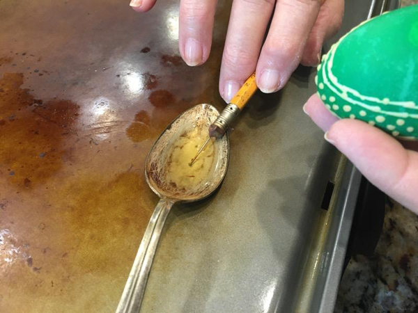 In the center of this shot, we see an large silver spoon with a rather battered and scratched bowl. It’s on a flat heated griddle, and contains a small amount of melted wax. Coming in from the top of the image, we see Barbara’s fingers holding a pencil lightly. A pin is stuck into the pencil’s eraser, and the head end of the pin is dipped into the melted wax. On the right edge of the image, we see a green egg that Barbara is going to apply the wax to.