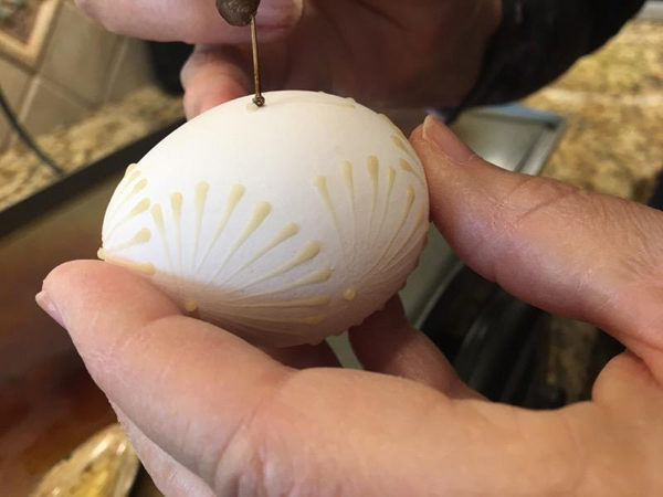 Closeup of Barbara drawing a traditional design on a white egg: teardrop-shaped lines radiating out from a central dot like the spokes of a bicycle wheel. She’s using her pin-on-a-pencil tool to apply melted wax in the “drop and pull” method.  A person holding the egg would be able to feel the wax lines, fatter at the outer edge of the circle and thinner as they go toward the dot at the center.  The beeswax is slightly golden on the white eggshell.