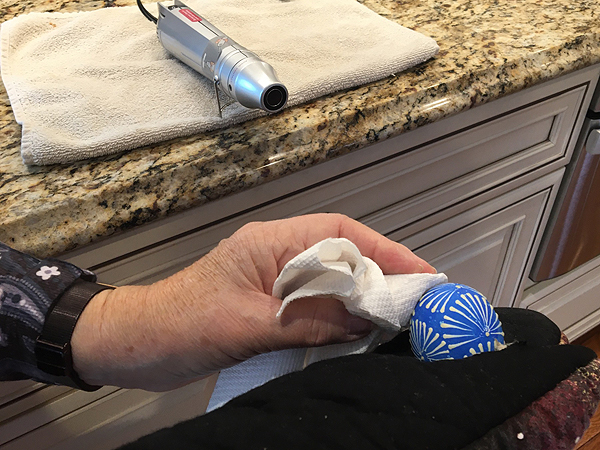 At the top of the image, an electric heat gun (which looks like a silver thermos bottle on its side) is blowing hot air toward a blue decorated egg, which Barbara is holding in a thick hot-mitt.  She is using a paper towel to wipe off the softened wax